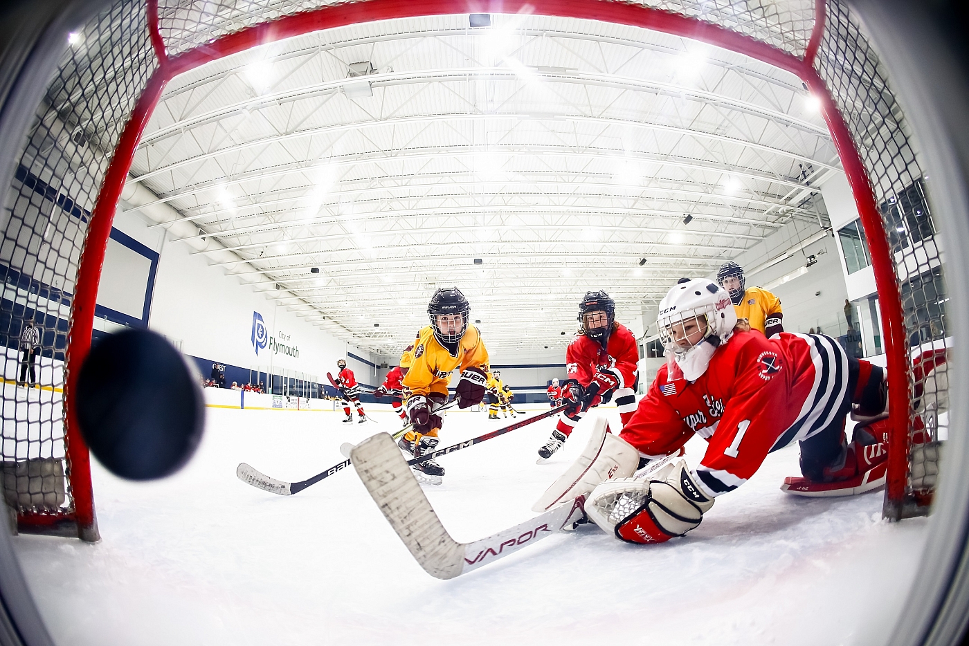 action photography during youth boys hockey game. Goal camera captures hockey puck flying into goal while both teams in red and yellow jerseys turn to try and fight for the puck.
